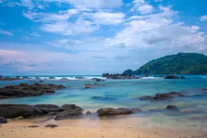 Idyllic tropical beach scene with rocky shoreline, clear blue waters, and lush hill in the background. jigsaw puzzle