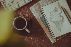 From above of sketchbook with person portrait near pen and pencil next to mug with drink on saucer with herbs on table near book and newspaper in light room jigsaw puzzle