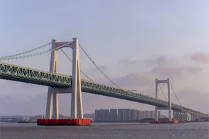 An expansive suspension bridge spanning across a serene river with urban skyline in the distance. jigsaw puzzle