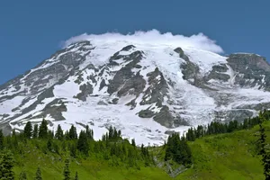 Snow-capped mountain peak with clouds, green trees, and grassy slopes. jigsaw puzzle