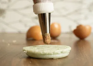 A piping bag nozzle icing a green macaron shell with eggshells in the background. jigsaw puzzle