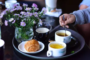 A hand stirring a cup of coffee next to a croissant and flowers on a breakfast tray. jigsaw puzzle