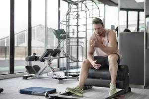 Tired young muscular male athlete in casual sportswear and sneakers sitting on bench and resting after intense weightlifting workout in modern fitness club jigsaw puzzle
