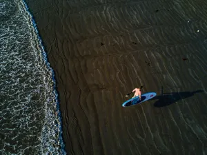 High-angle aerial shot of a surfer carrying a surfboard on a sandy beach by the sea. jigsaw puzzle