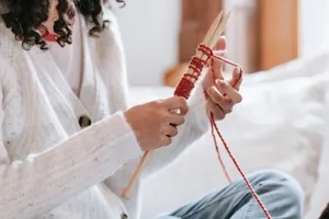 Crop unrecognizable female in casual wear knitting with needles while sitting on bed with cushion in light room at home jigsaw puzzle