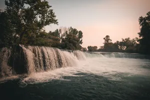 A gentle waterfall cascading under a clear sky surrounded by lush green trees at dusk. jigsaw puzzle