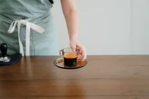 Crop unrecognizable female barista in light green apron passing hot aromatic espresso in stylish transparent cup above wooden table jigsaw puzzle