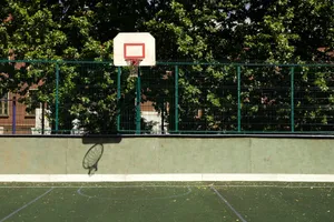 A sunlit outdoor basketball hoop with green fence and court surface. jigsaw puzzle