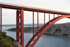 A striking red arched bridge spans a tranquil body of water surrounded by rocky hillsides. jigsaw puzzle
