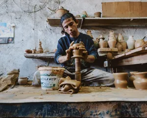 A potter shapes clay on a wheel in a rustic studio surrounded by finished pottery pieces. jigsaw puzzle